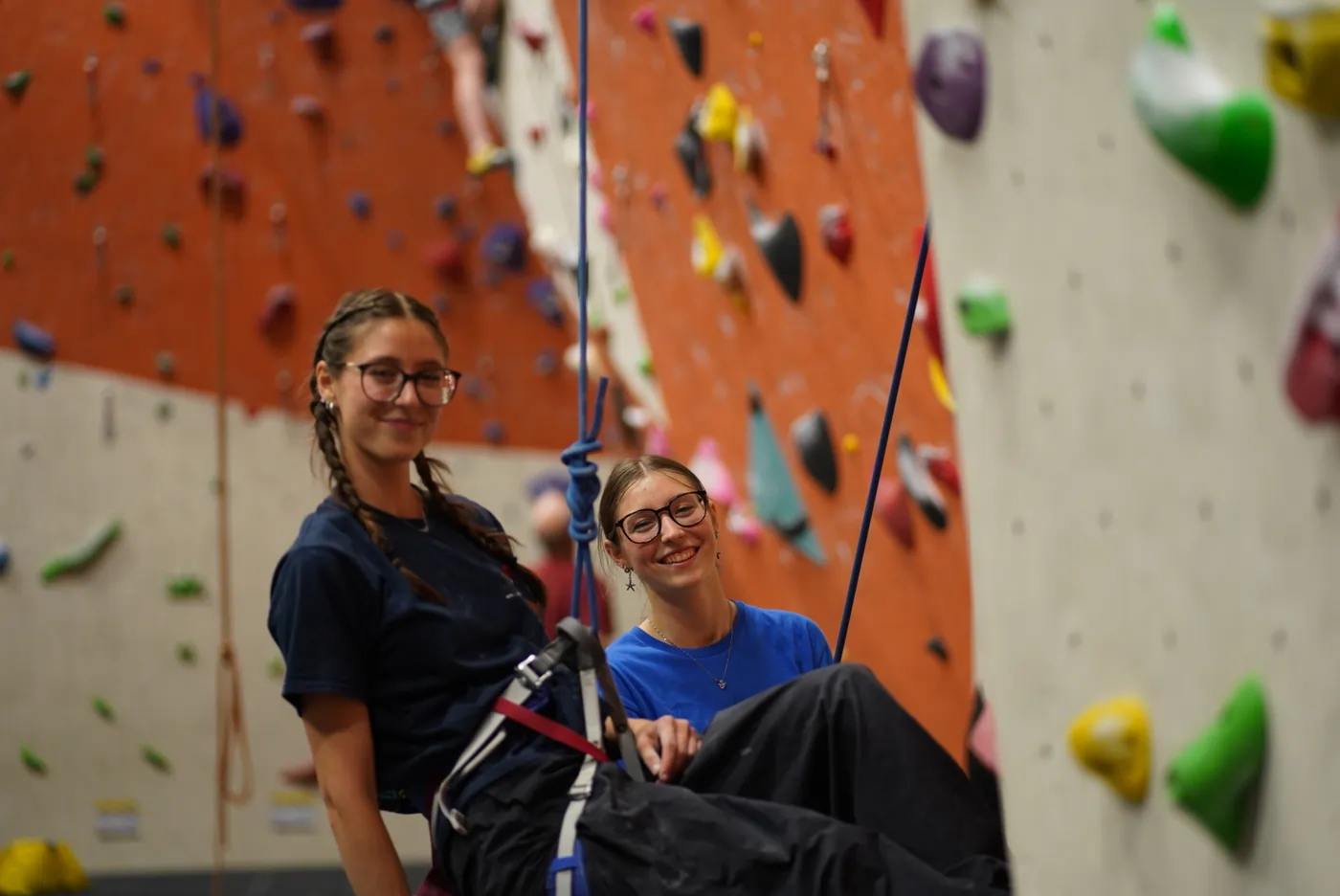 Two female climbers face towards the camera smiling, with one still hanging from their harness on the wall at Dublin Climbing Centre