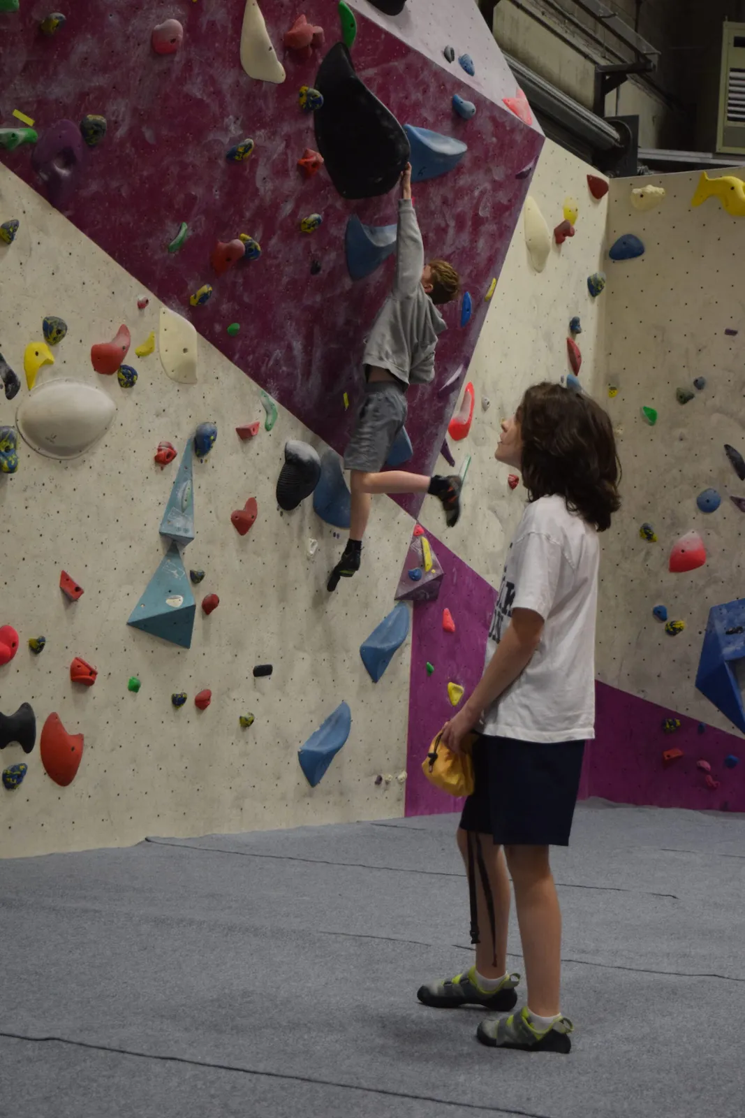 One child scales the bouldering wall while another prepares to climb and studies a route
